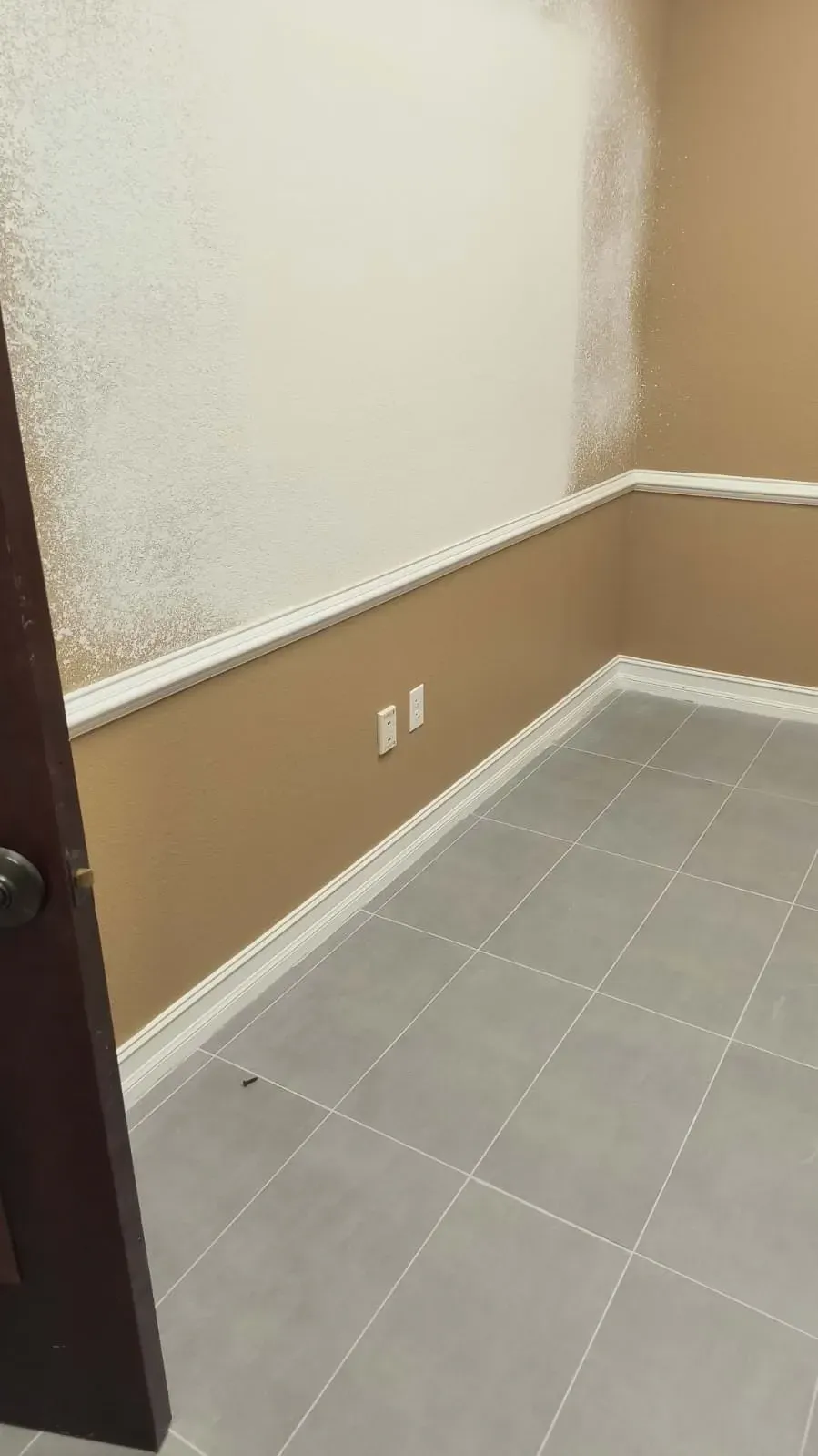 Empty room with tan walls, white molding, and gray tile floor. One wall is partially painted white.