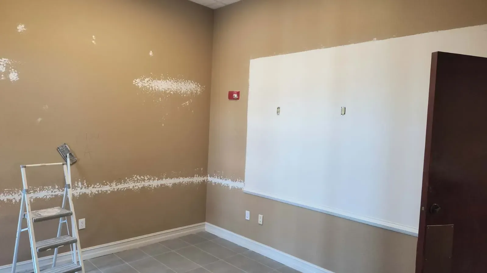 Room interior, partially painted. Beige walls, white patch repairs, ladder, white painted section, brown door.