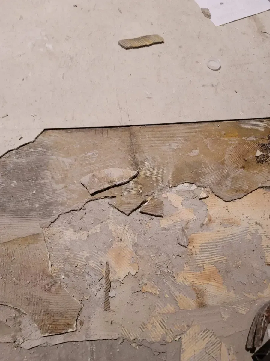 Damaged floor with broken tiles and exposed subfloor; light brown and beige tones.