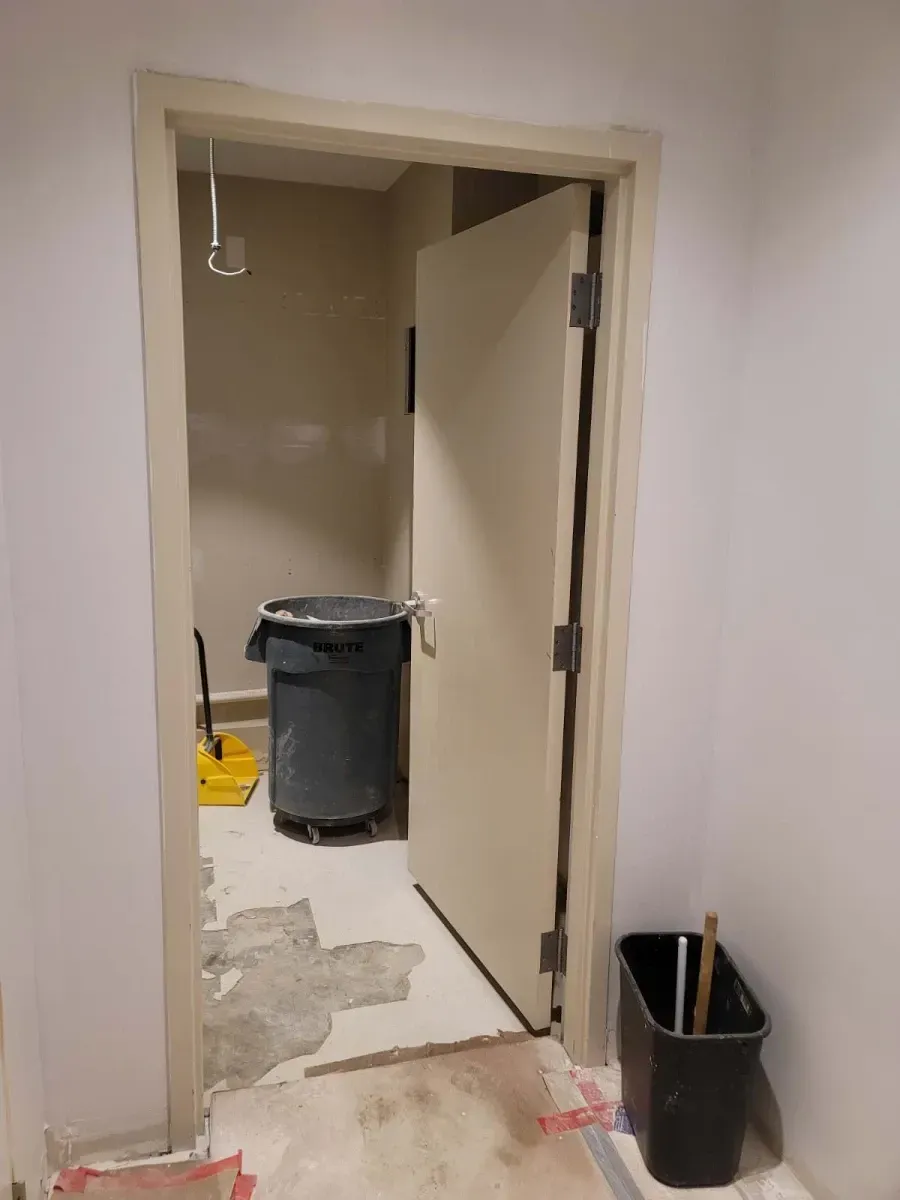 Open doorway leads to a small room with trash can and debris. Pale walls and door frame.