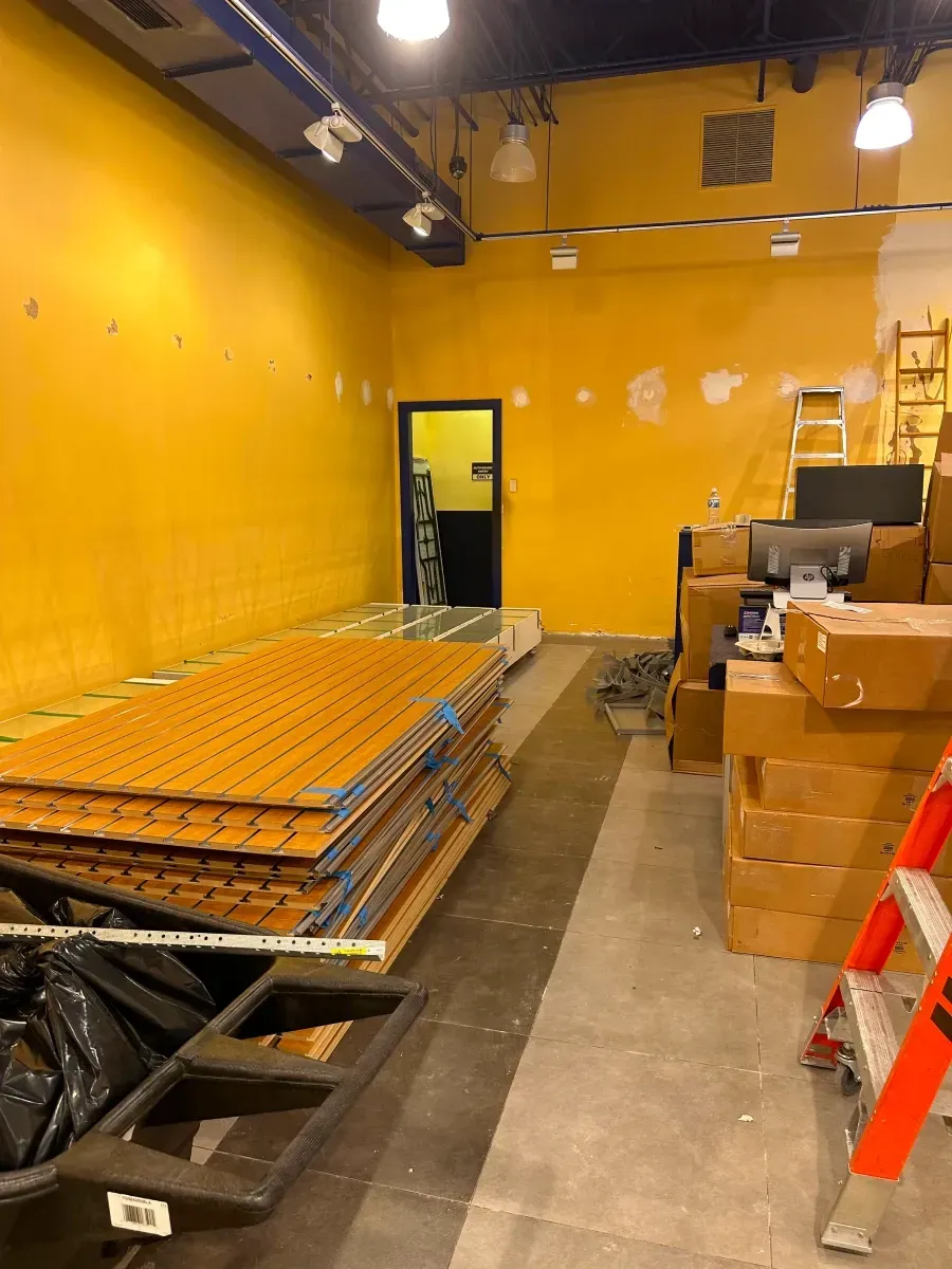 Interior of a yellow-painted store under construction. Stacked panels, boxes, ladder, and a door are visible.
