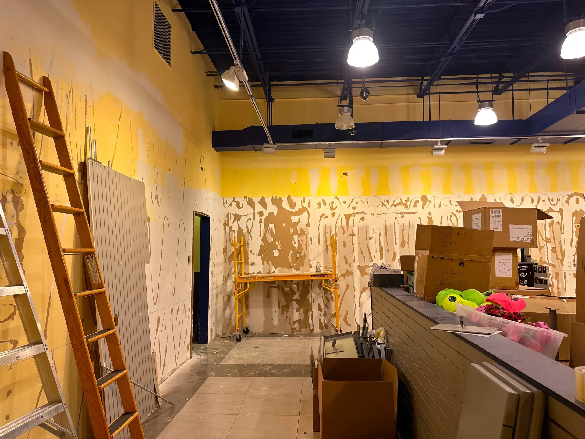 Interior construction in progress, with exposed wall, ladder, scaffolding, boxes, and overhead lights.