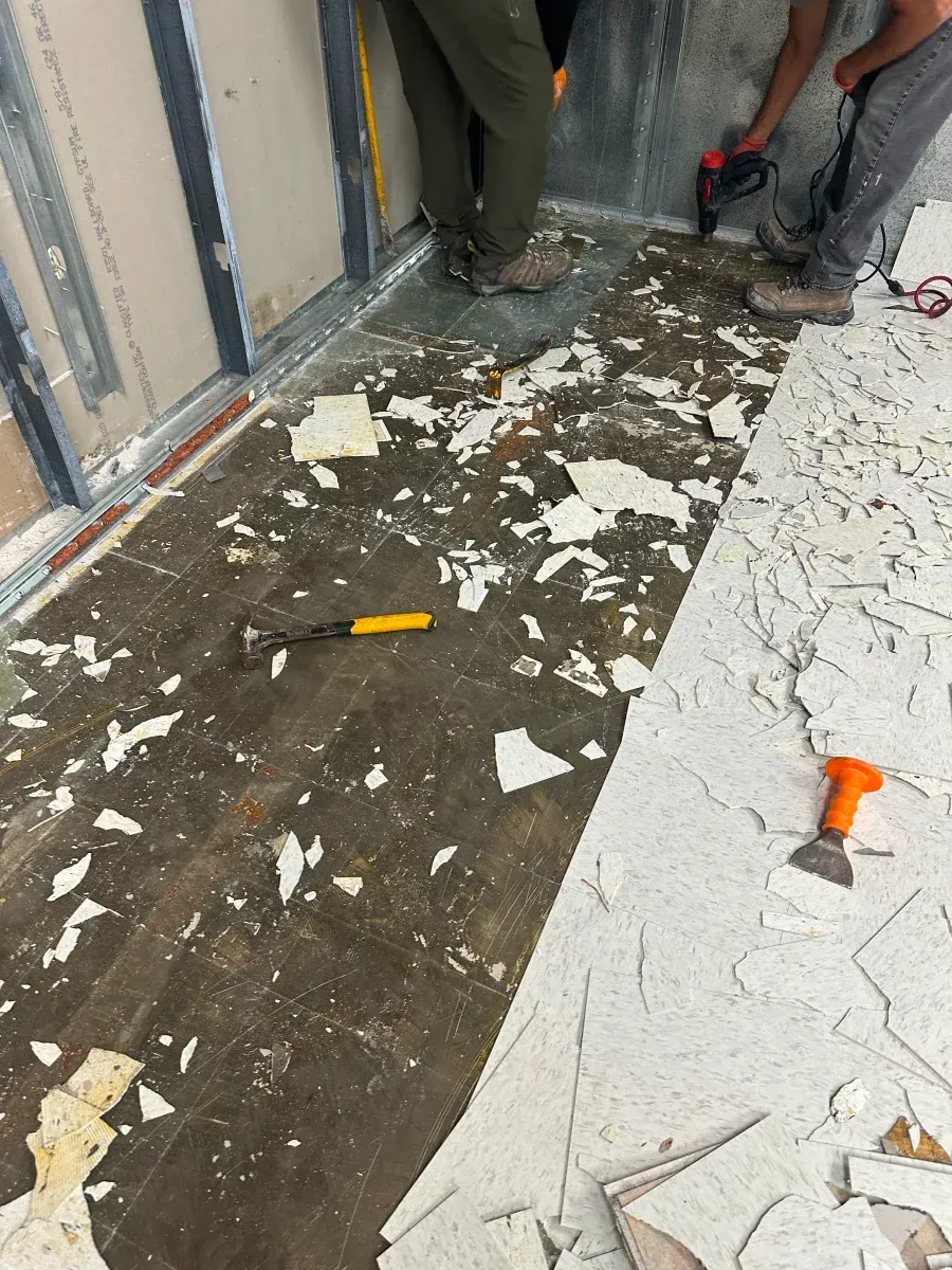 Workers removing flooring in a room. Debris covers the floor. One worker uses a tool.