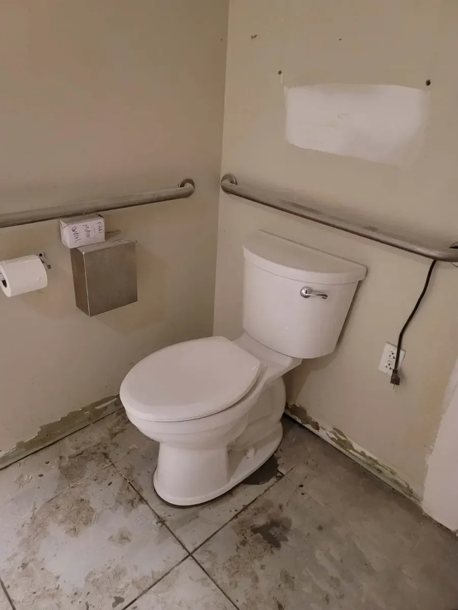White toilet in corner of a bathroom with grab bars and paper towel dispenser.  Dirty floor.