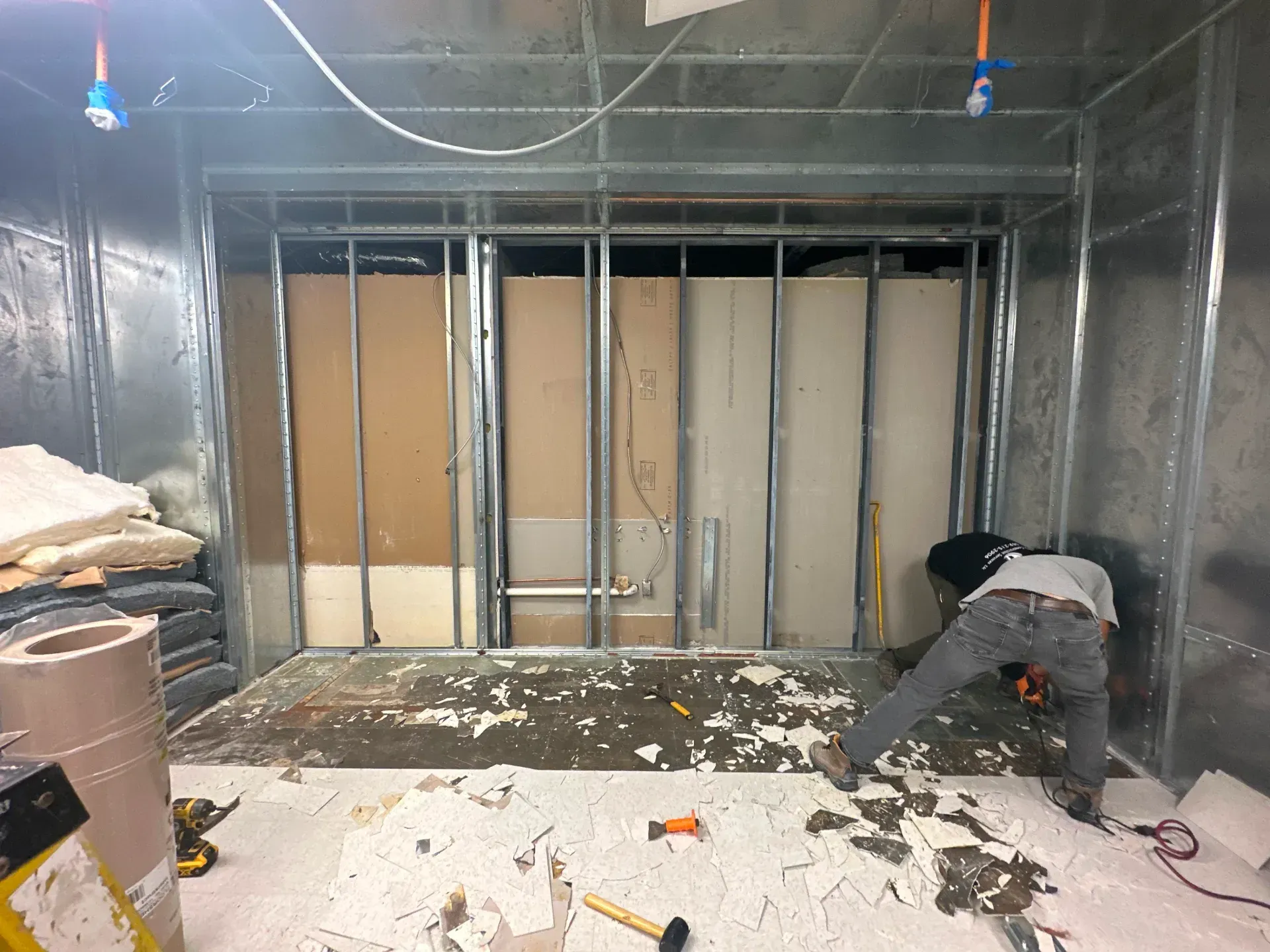 Construction worker removing debris from a room with metal stud framing and partially installed drywall.