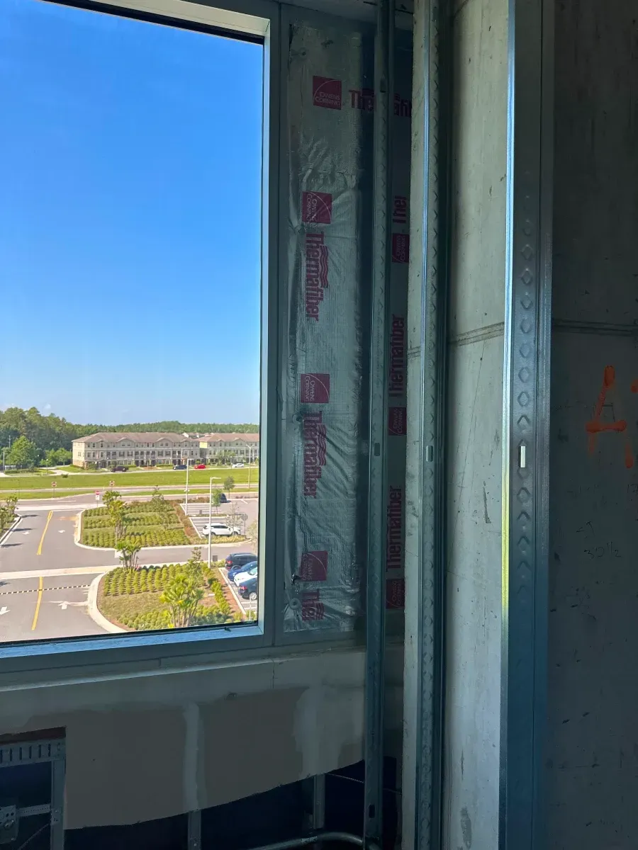 Window view of a building under construction, showing framing, vapor barrier, and exterior landscape on a sunny day.