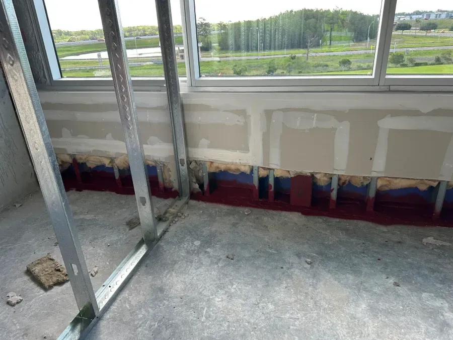Interior wall under construction with metal framing, drywall, and insulation visible. Concrete floor, window in background.