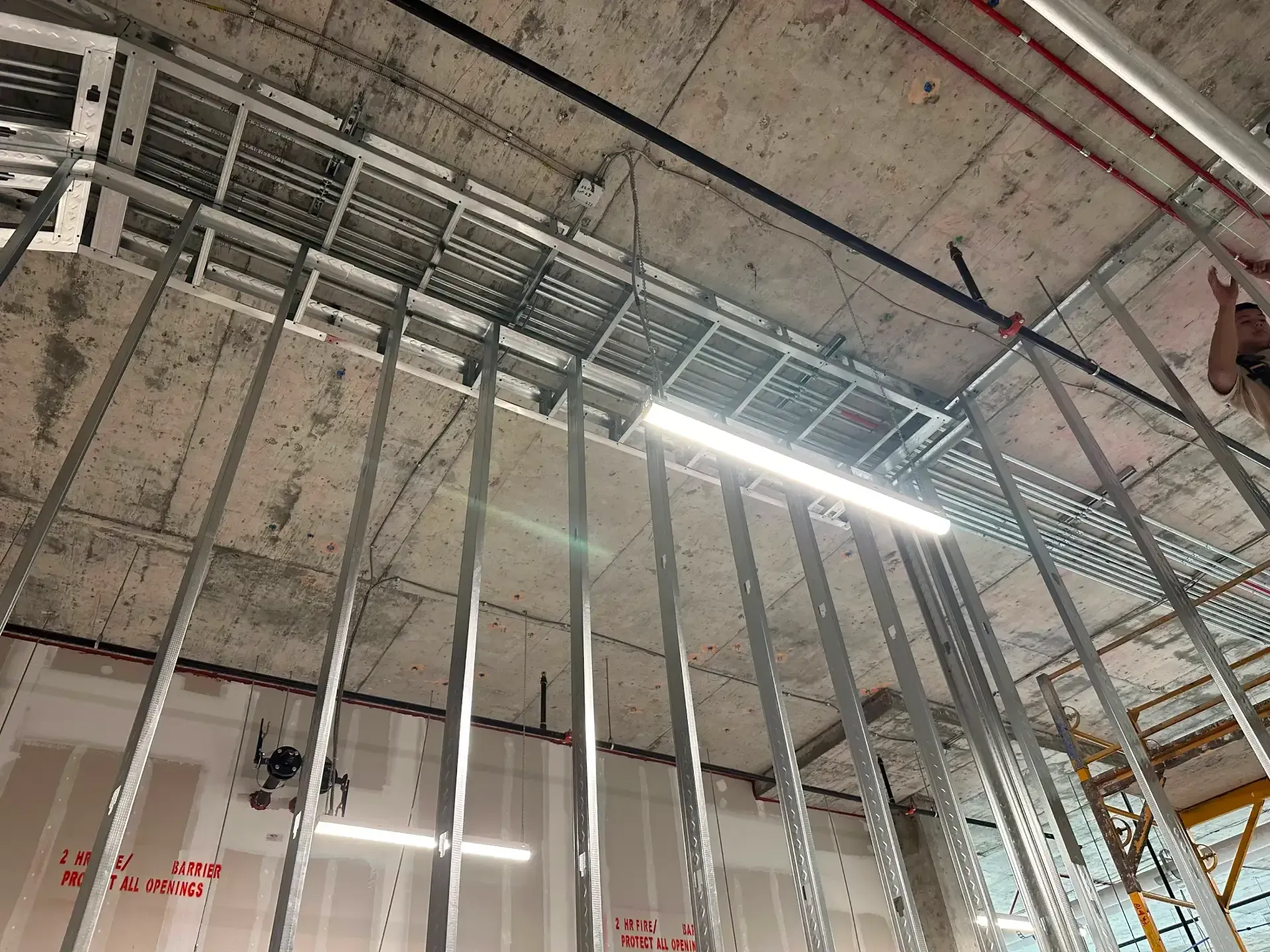 Interior of a building under construction. Metal studs frame a wall; ceiling contains conduit and lights.