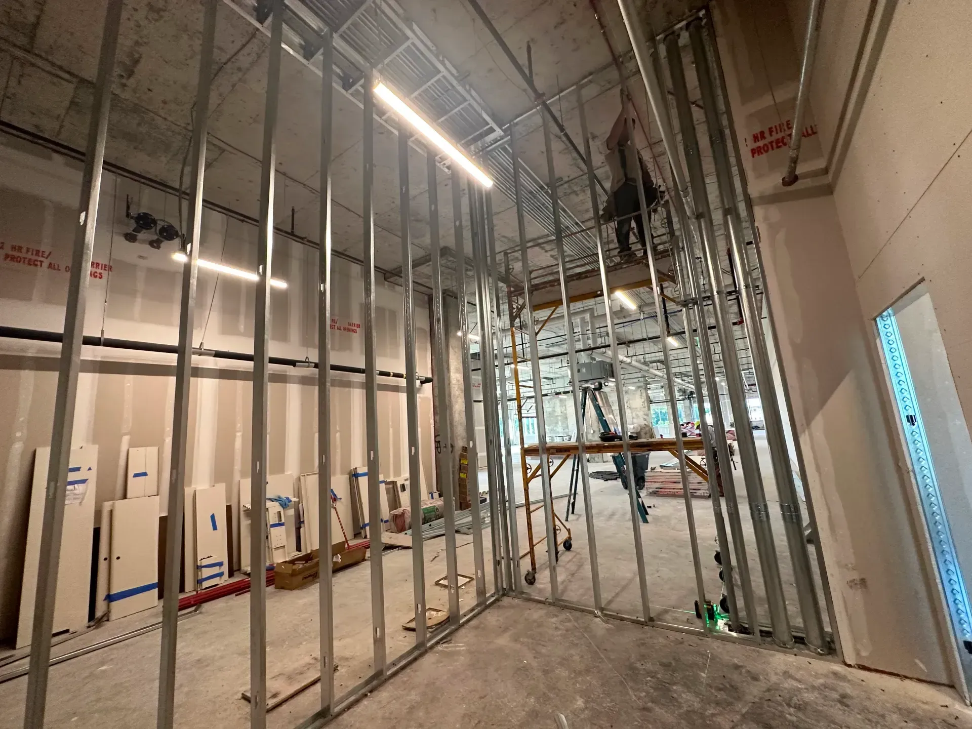 Interior construction site with metal studs forming walls. A worker stands on scaffolding in the distance.