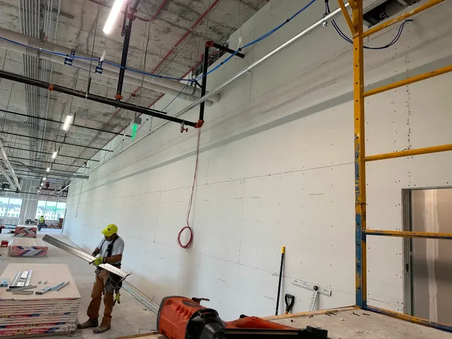 Construction worker installing drywall in a building. Scaffolding, tools, and pipes visible.