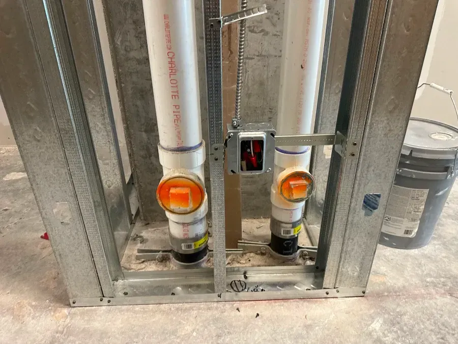 Plumbing pipes within a metal frame, set in a construction zone. Orange caps and an electrical box are visible.