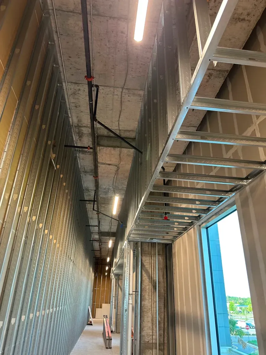 Interior hallway under construction with metal framing, exposed pipes, and a window.
