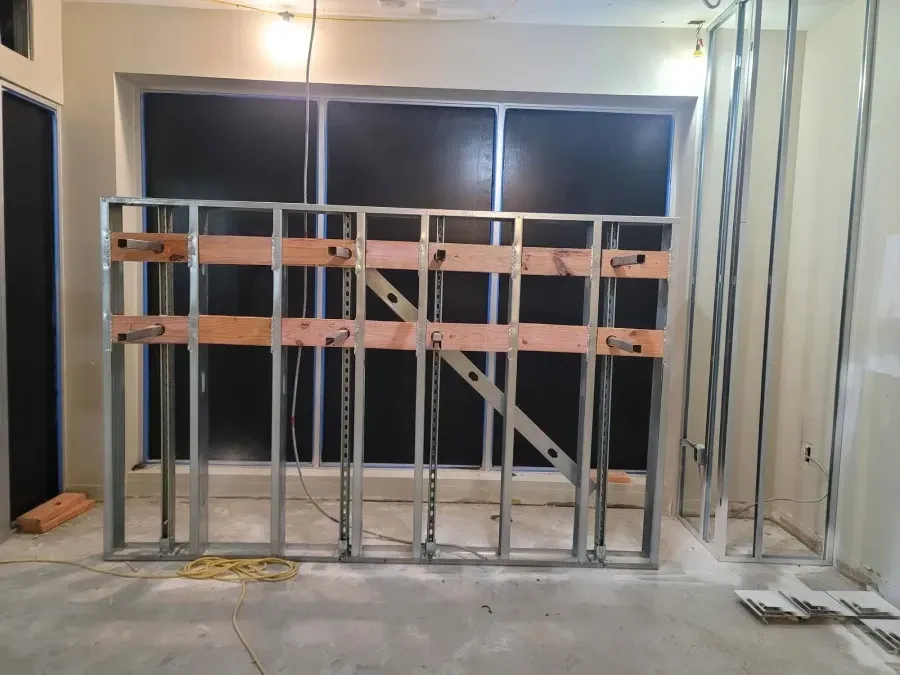 Metal and wood framing of a wall with pipes running through it, in a room with a window.