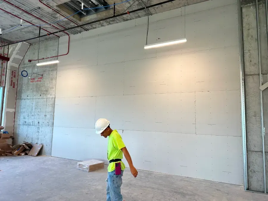 Construction worker in hard hat, examining drywall. Interior, unfinished space, fluorescent lights.