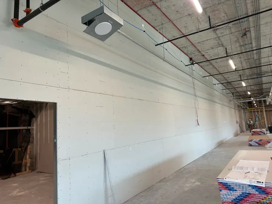 Interior of a construction site with drywall on a long wall, exposed pipes and ceiling lights.