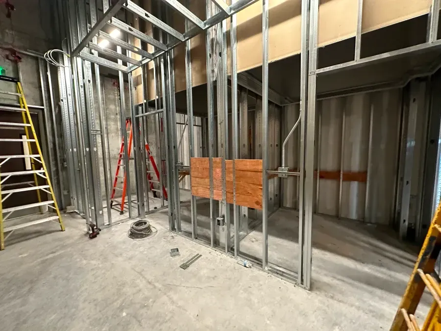 Construction site with metal framing.  Ladder, orange and red ladders, electrical wiring, and a wooden panel are visible.
