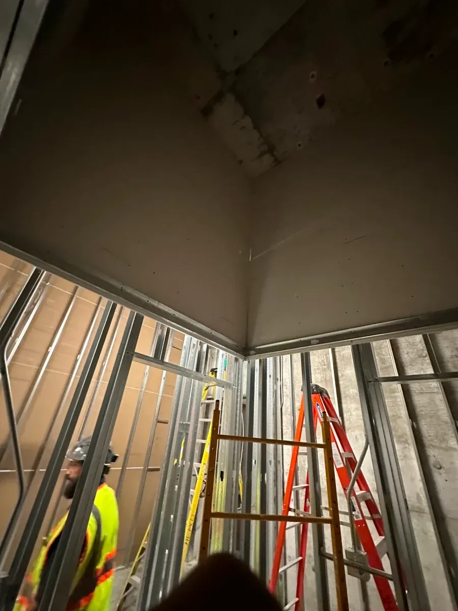 Construction worker in a high-vis vest stands near metal framing and two ladders, working on a corner ceiling.