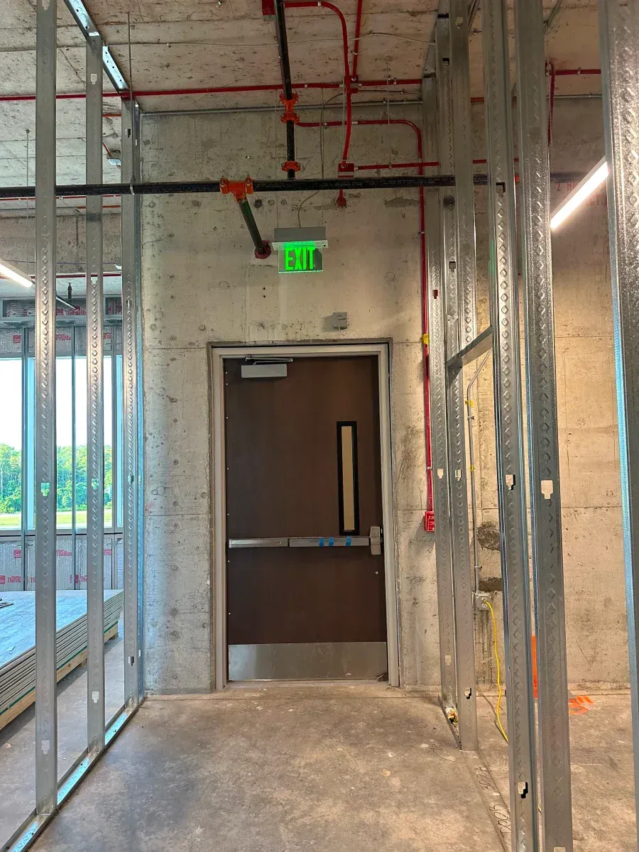 Hallway with metal studs, a door with exit sign, and fire sprinkler pipes.
