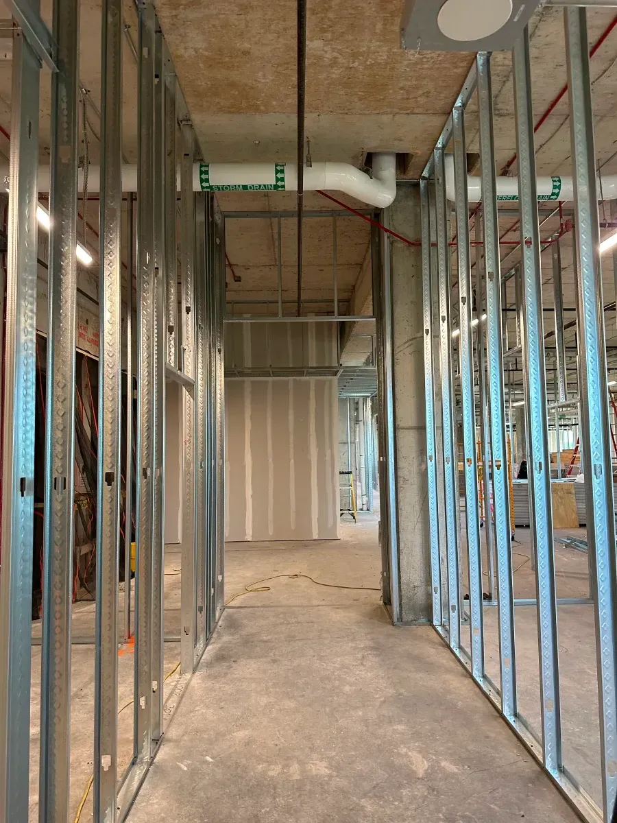 Interior view of a construction site with metal framing. A corridor leads to a wall. Piping and wiring are visible overhead.