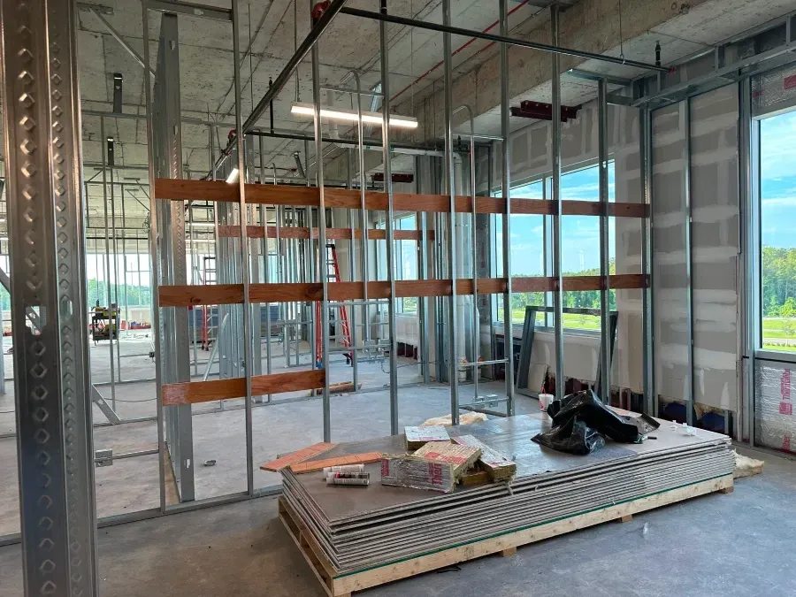 Interior of a building under construction, metal framing and drywall. Construction materials on the floor near large windows.