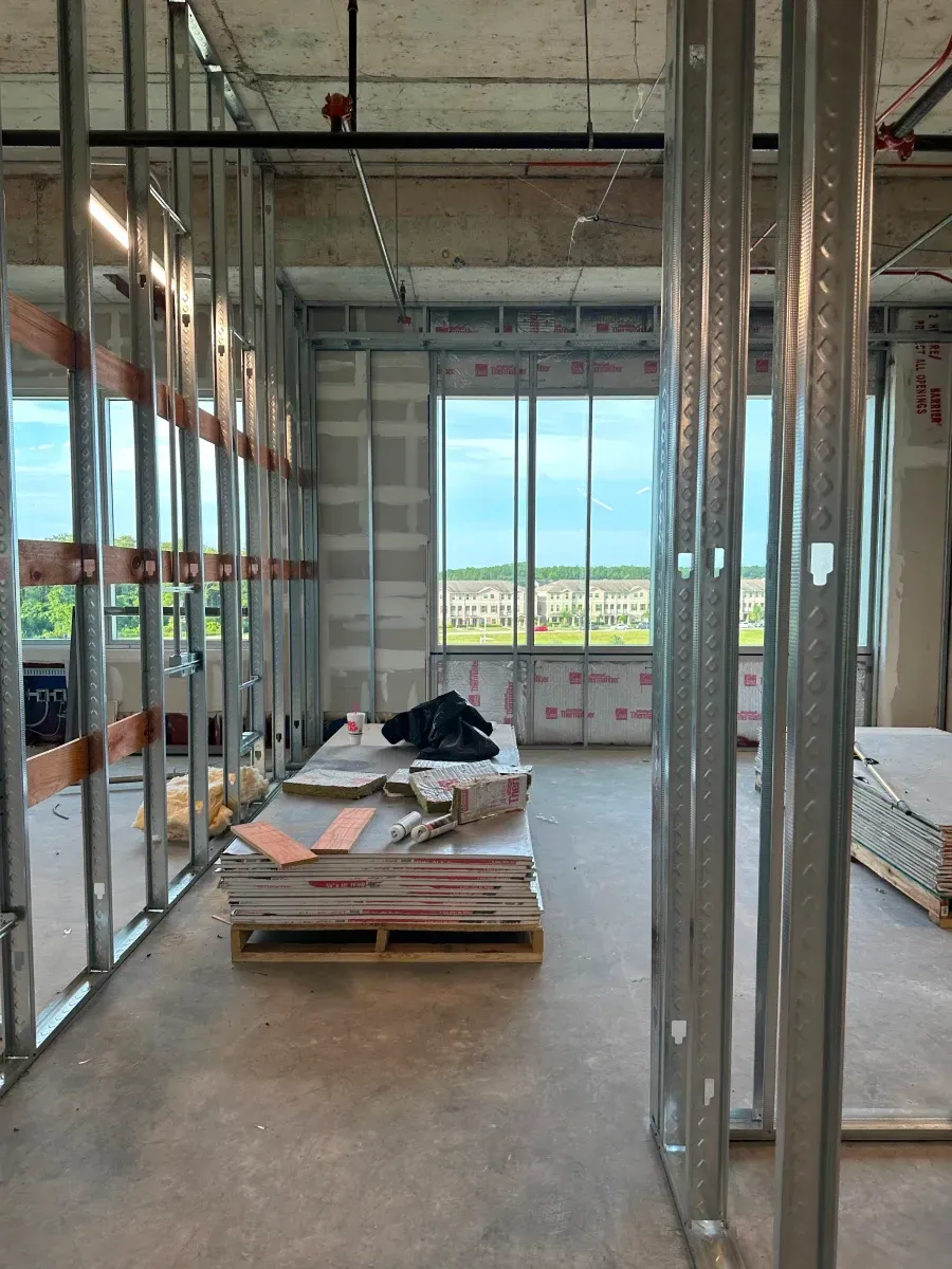 Interior construction site with metal studs, drywall, and a window overlooking a landscape.
