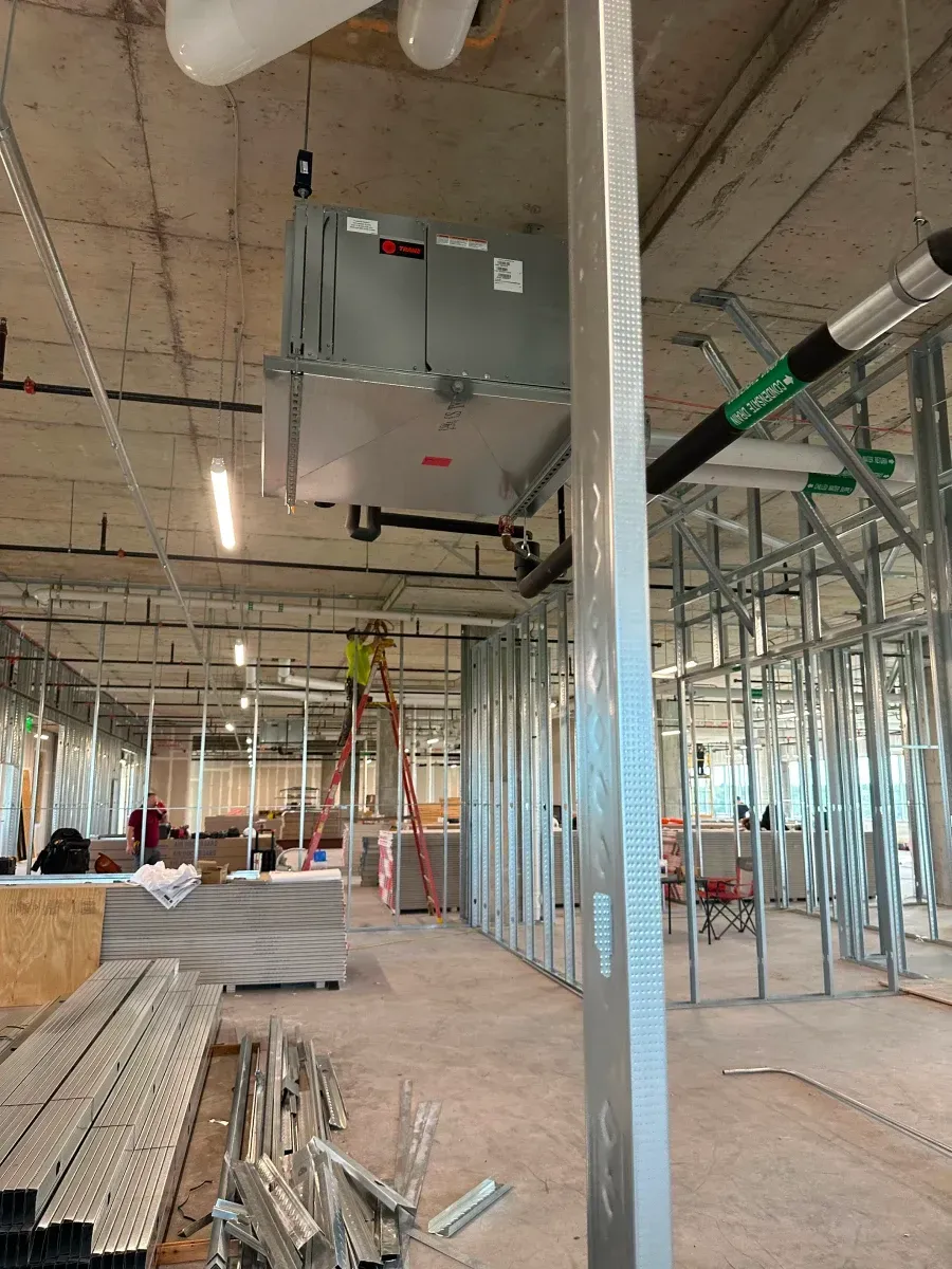 Construction site: metal studs, HVAC unit, worker on ladder, exposed ceiling, unfinished walls.
