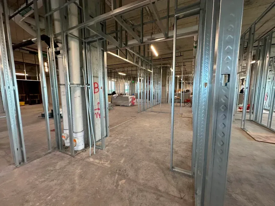 Interior construction site, metal studs framing walls and ceiling. Concrete floor, pipes visible.