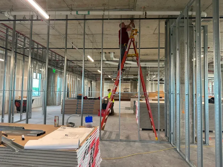 Construction workers inside a building under renovation; one on a ladder working on ceiling fixtures.