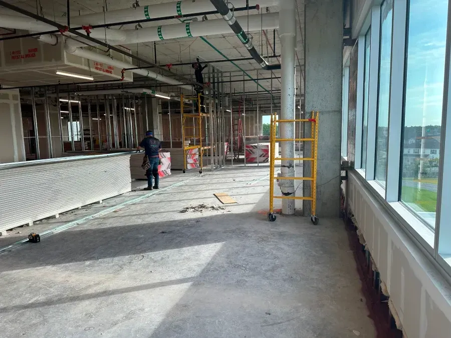 Construction site with exposed framing, pipes, and scaffolding; a worker installs drywall.