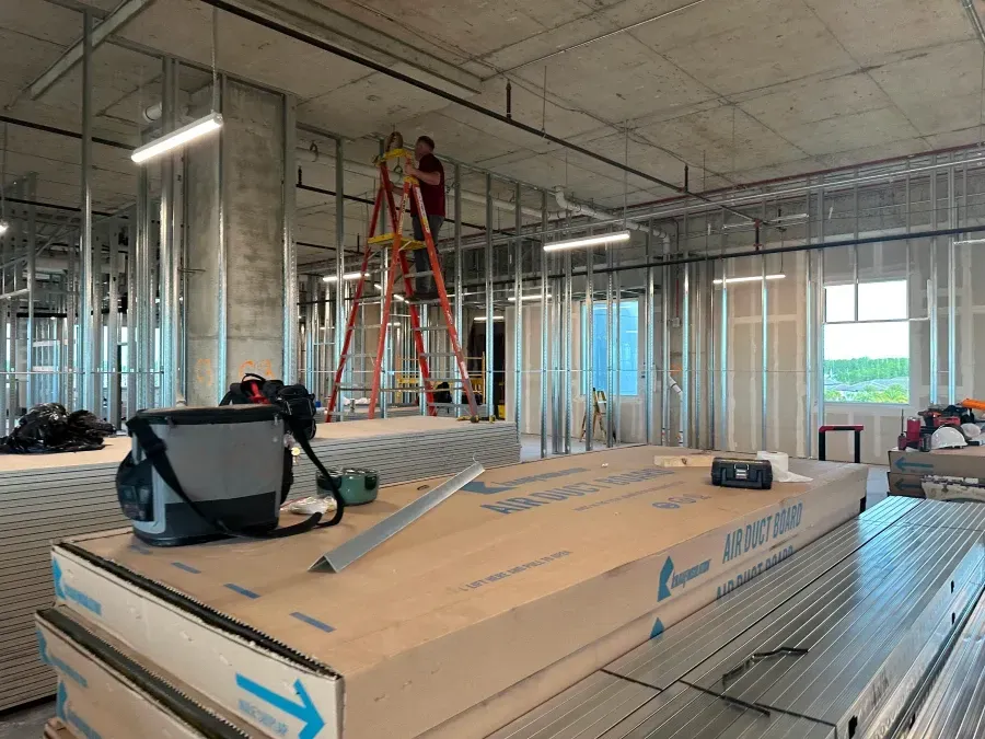 Construction site: metal studs, drywall, person on ladder, materials stacked. Interior view.
