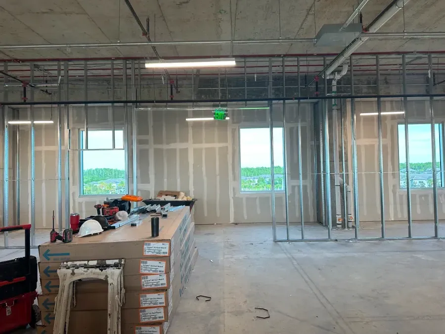 Interior view of a construction site with metal framing, drywall, windows, tools, and stacked materials.