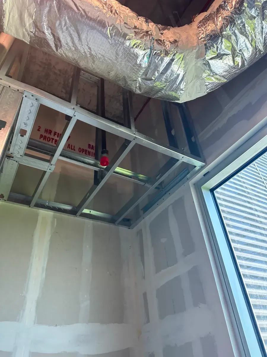 Interior view of construction: metal framing for a ceiling with exposed ductwork, drywall walls, and a window.