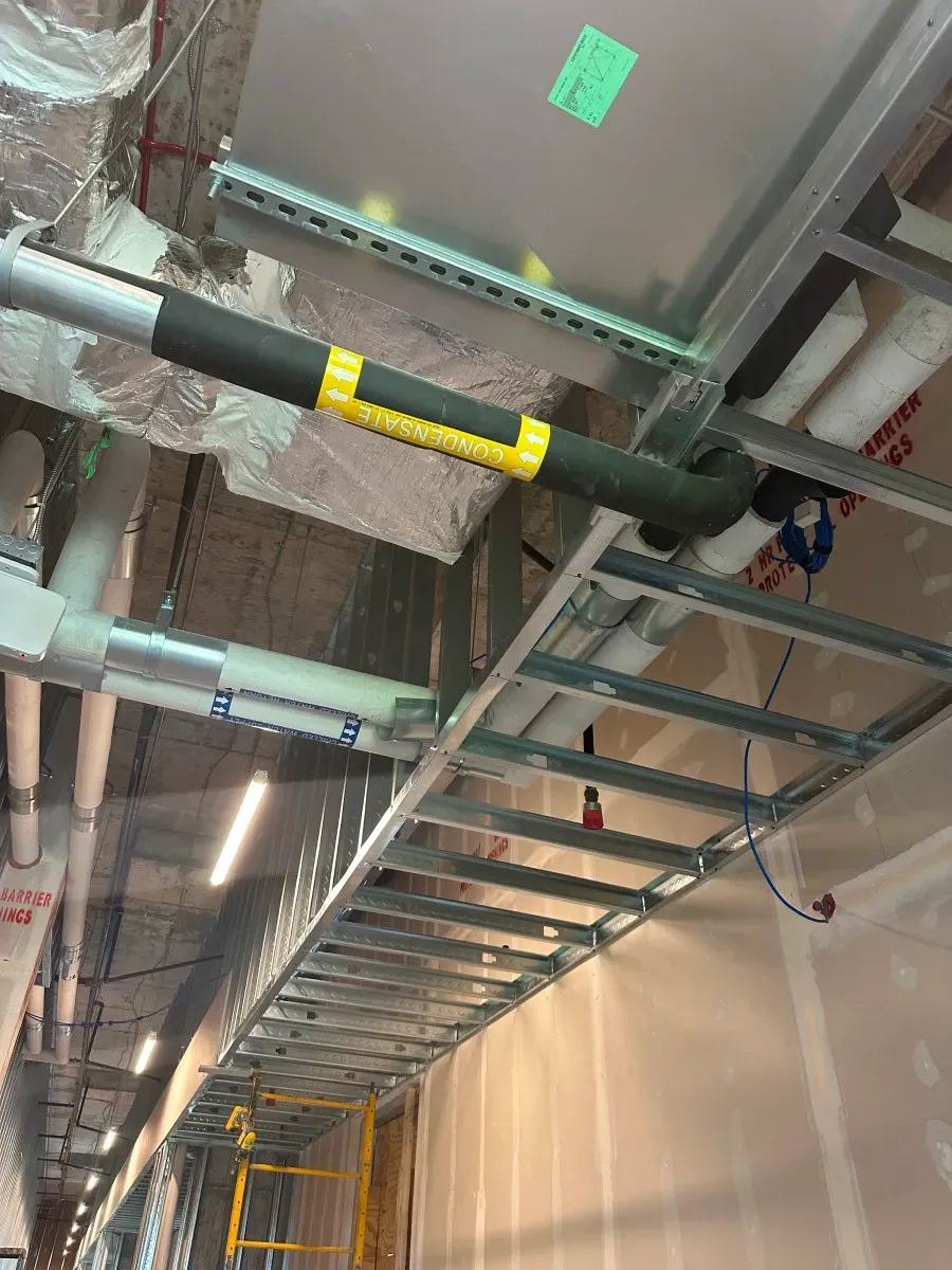 Construction site ceiling with pipes, cable trays, and framing. Insulation and wiring visible.