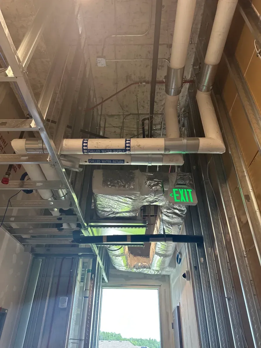 Construction of a narrow corridor with metal framing, pipes, ductwork, and an exit sign.