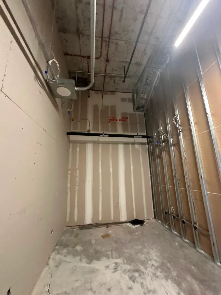 Construction site interior with drywall, metal studs, electrical conduit, and exposed concrete ceiling.