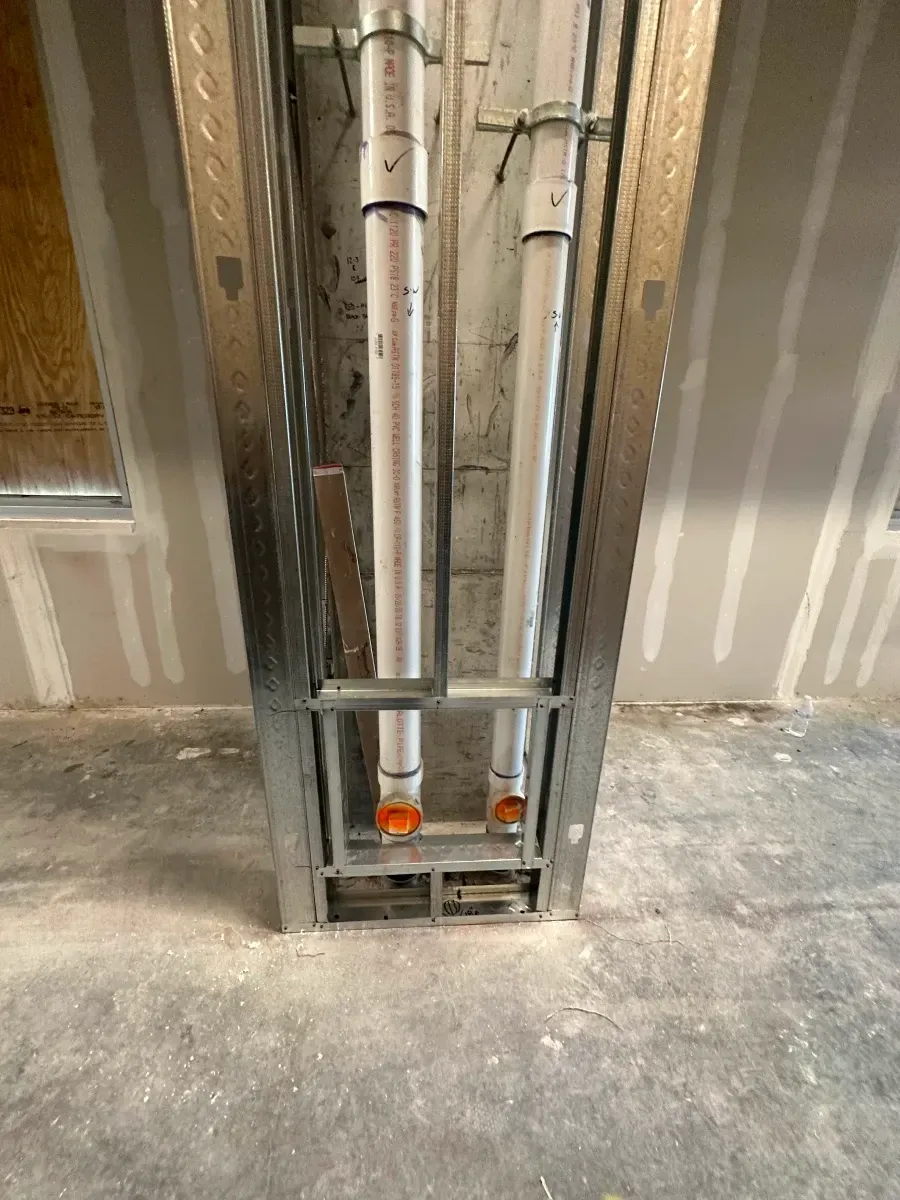 Metal framing around plumbing pipes in a construction setting. Gray pipes, orange fittings, and concrete floor.