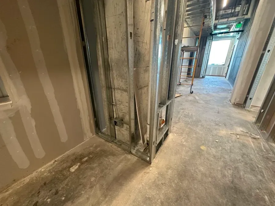 Hallway under construction; metal framing, drywall, concrete floor, doorway with light.
