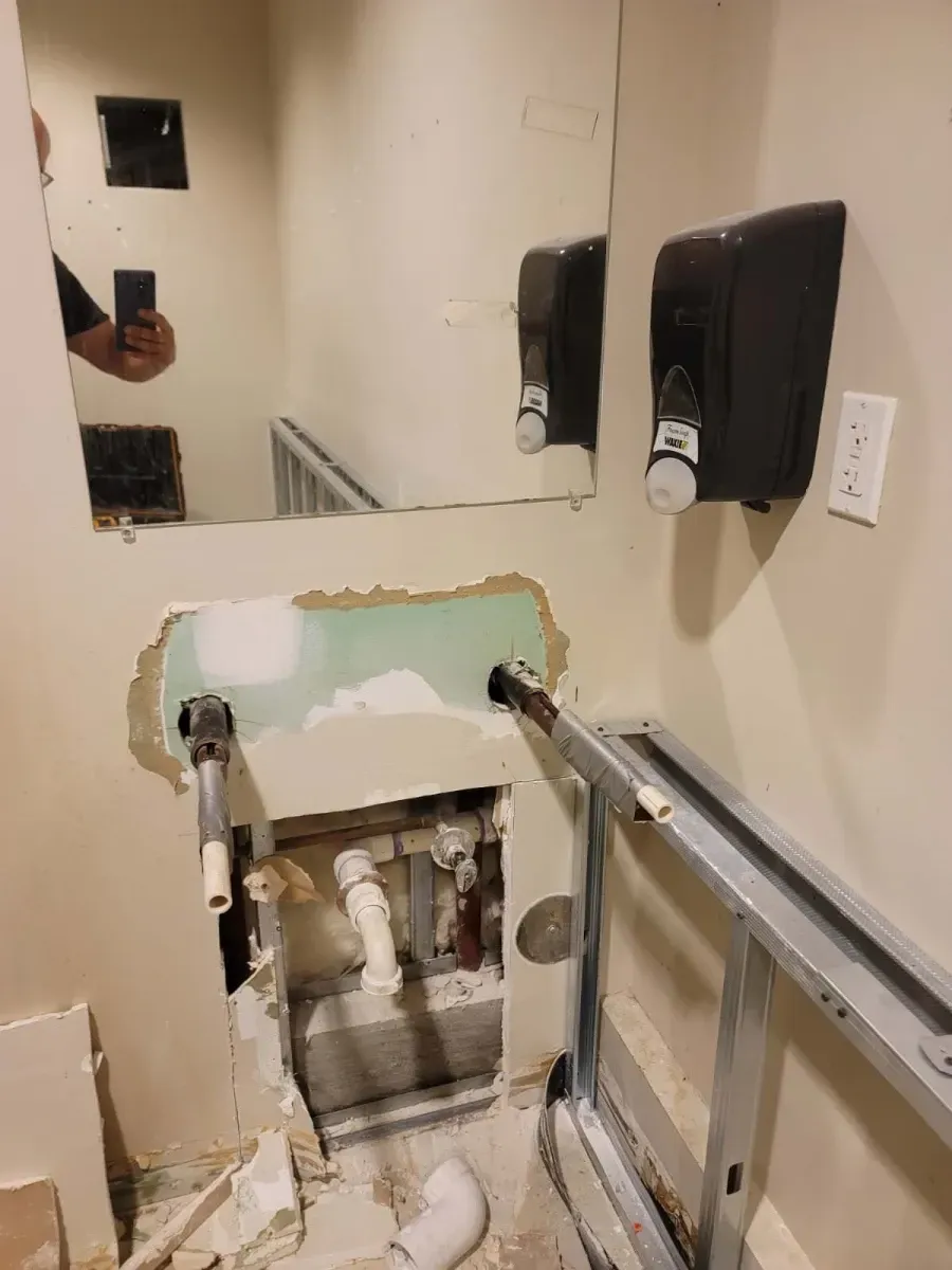 Bathroom wall under construction, exposed plumbing, mirror, soap dispensers. Metal framing, drywall removed.