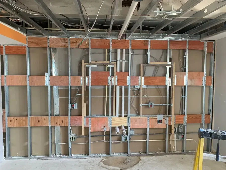Wall under construction, exposed metal studs, wooden support beams, electrical wiring and plumbing visible.