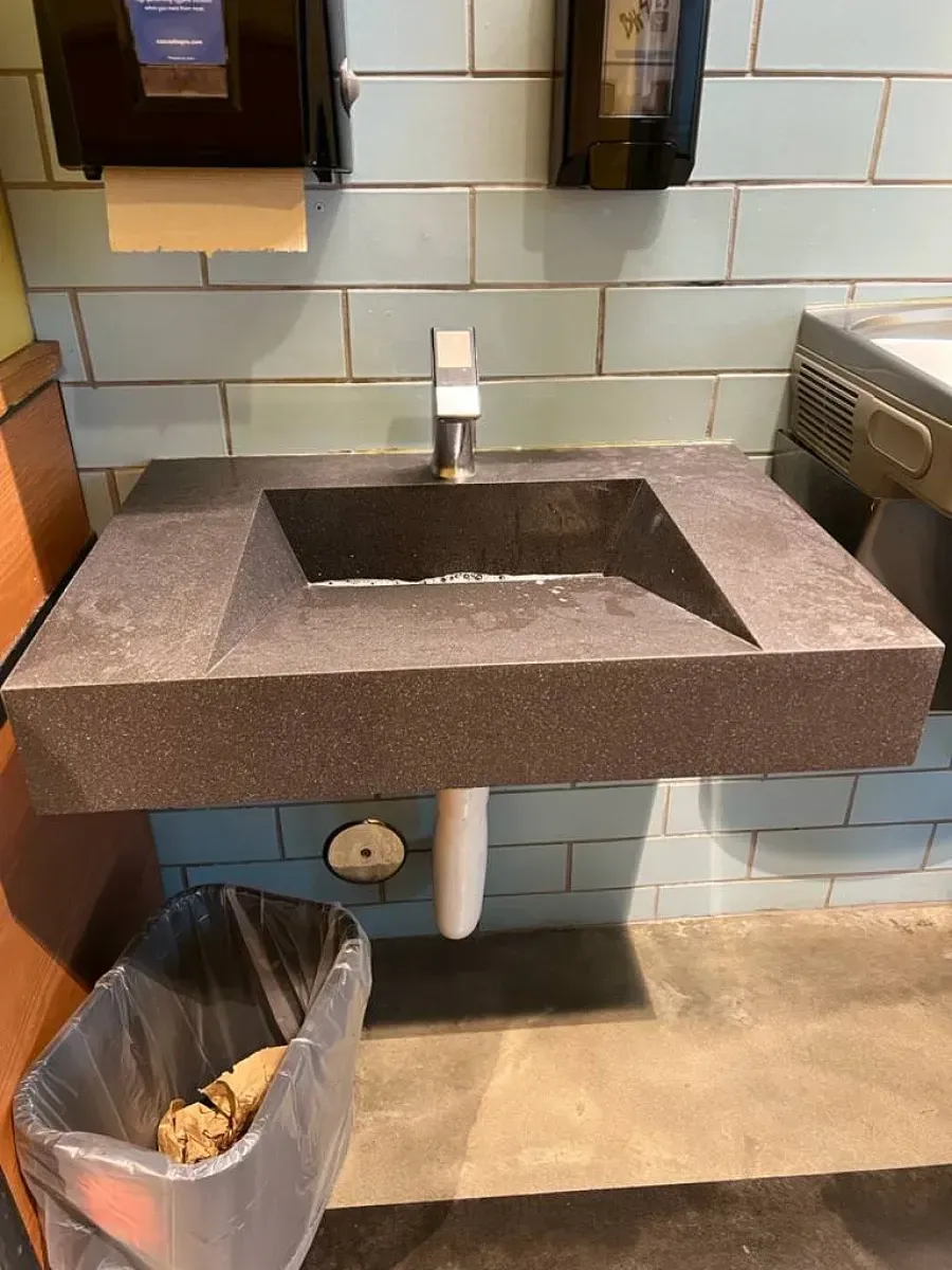 A dark gray, rectangular stone sink with a chrome faucet. Blue tiled wall in the background. A trash can is on the floor.
