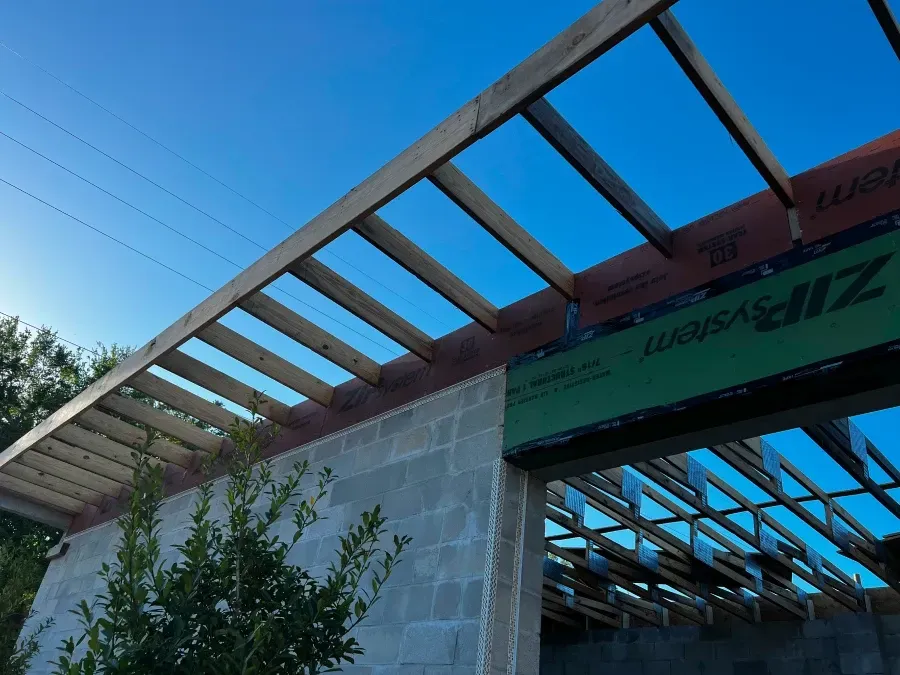 Construction of a building roof with wooden beams, cinder block walls, and blue sky background.