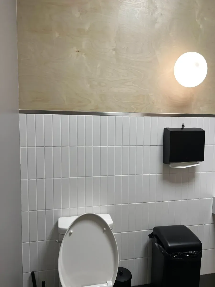 Bathroom with white tile, a toilet, a trash can, and a round wall light.