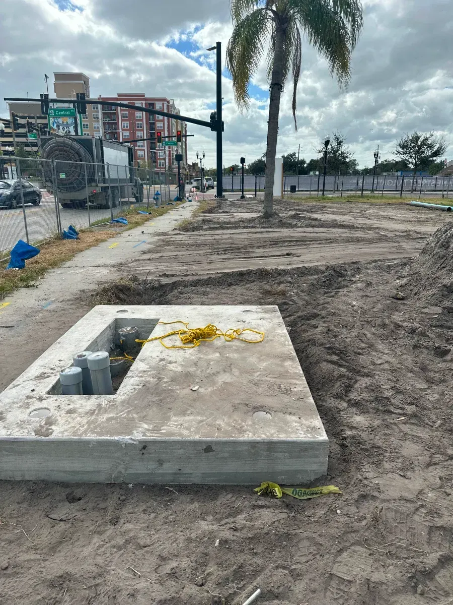 Construction site with concrete box, dirt, sidewalk, street, palm tree, and buildings.