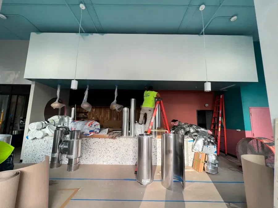 Construction site: Worker on ladder, installing fixtures above a counter, various equipment around.