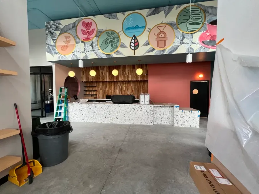 Interior view of a cafe under construction with colorful mural, counter, and equipment; exposed wires and debris.
