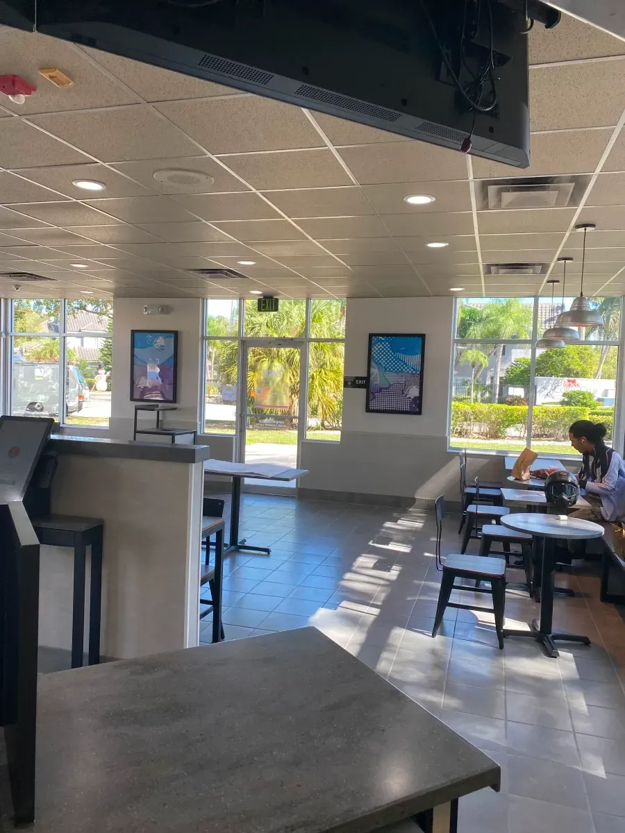 Interior of a bright cafe with tables and chairs near windows and a door. Two posters on walls.