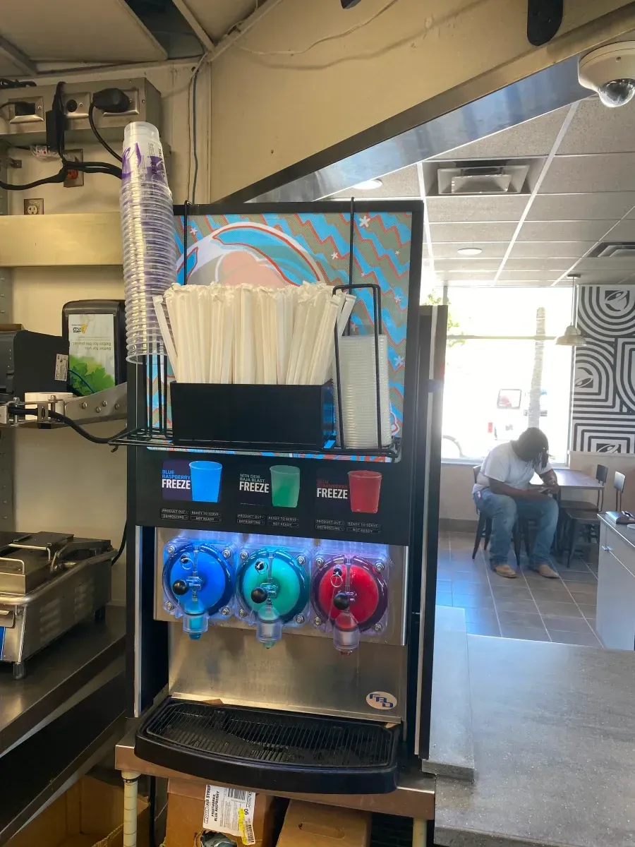Slushie machine with three flavors, cups, straws, and a person seated nearby in a restaurant.