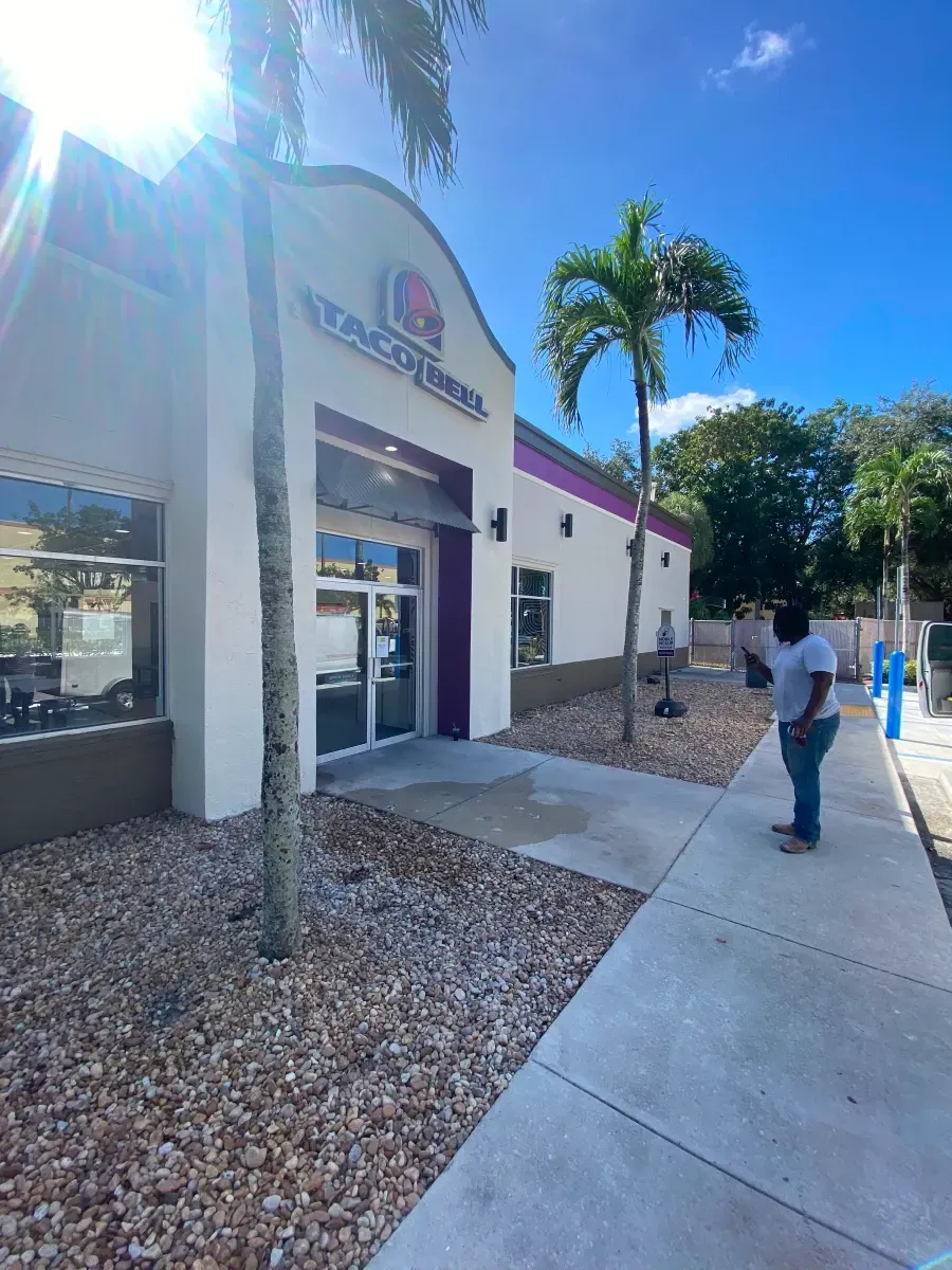 Taco Bell restaurant exterior; person stands on sidewalk. Building is white with purple accents. Trees and blue sky.