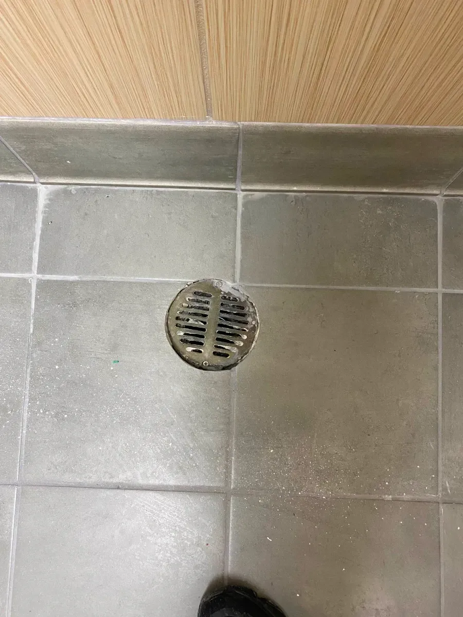 Shower floor with drain. Gray tiles, light wood paneling, and a foot are visible.