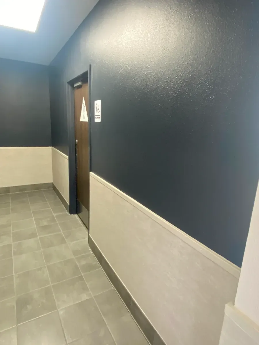 Dark blue painted hallway with beige wainscoting and a closed brown door. Gray tiled floor.